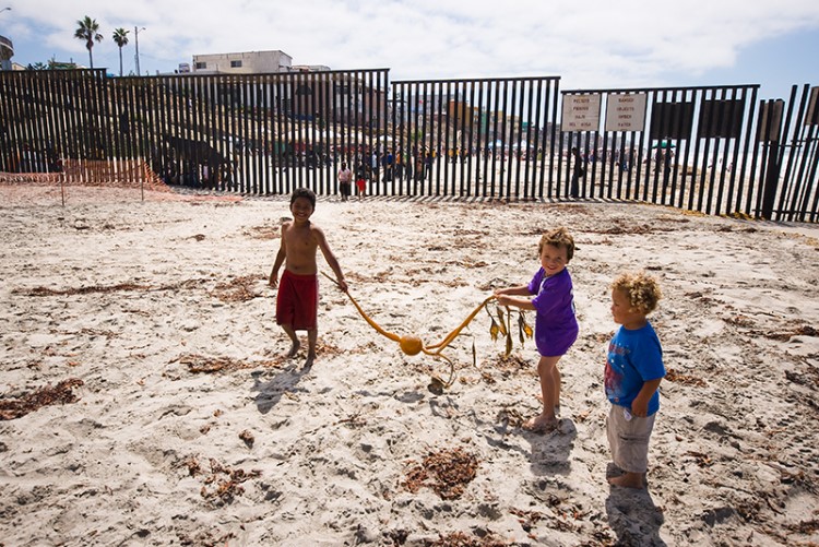 children play with kelp wrack on the beach in front of border wall 